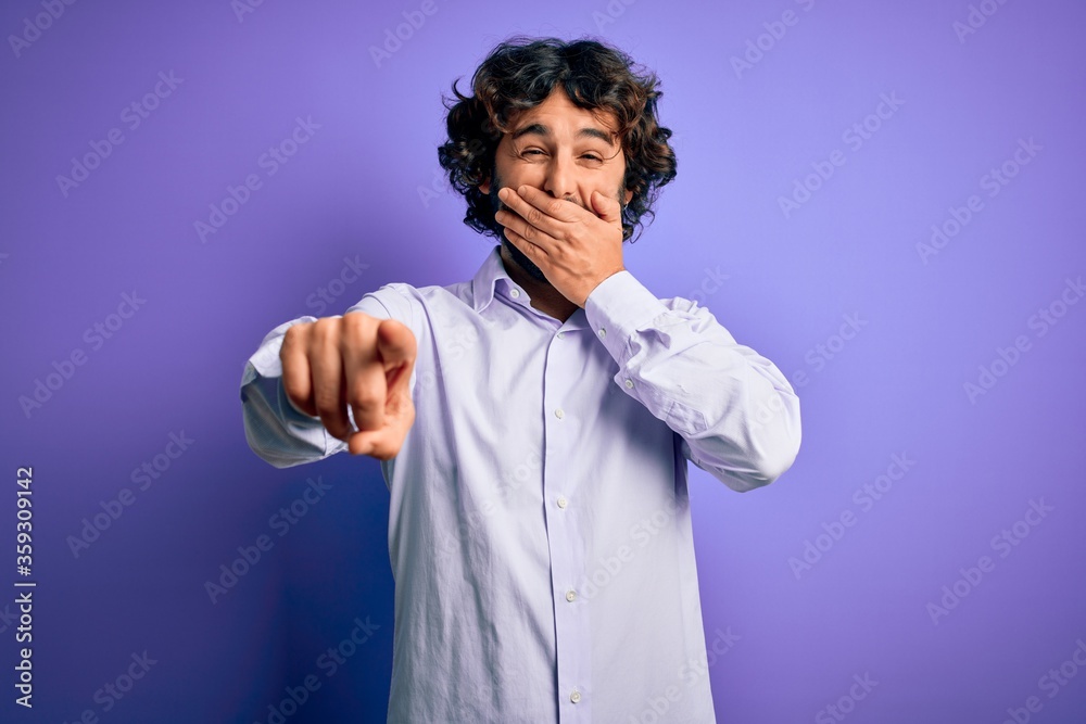 Young handsome business man with beard wearing shirt standing over purple background laughing at you, pointing finger to the camera with hand over mouth, shame expression