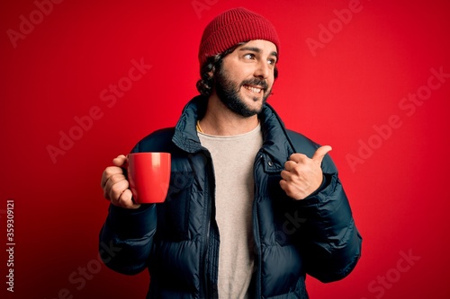 Young handsome man with beard wearing coat drinking cup of hot coffee over red background pointing and showing with thumb up to the side with happy face smiling
