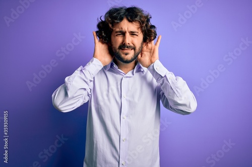 Young handsome business man with beard wearing shirt standing over purple background Trying to hear both hands on ear gesture, curious for gossip. Hearing problem, deaf © Krakenimages.com
