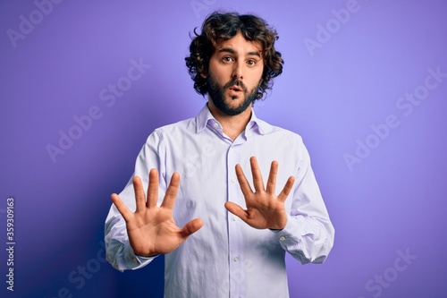 Young handsome business man with beard wearing shirt standing over purple background Moving away hands palms showing refusal and denial with afraid and disgusting expression. Stop and forbidden.