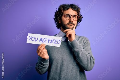 Handsome business man with beard holding you are fired message over purple background serious face thinking about question, very confused idea
