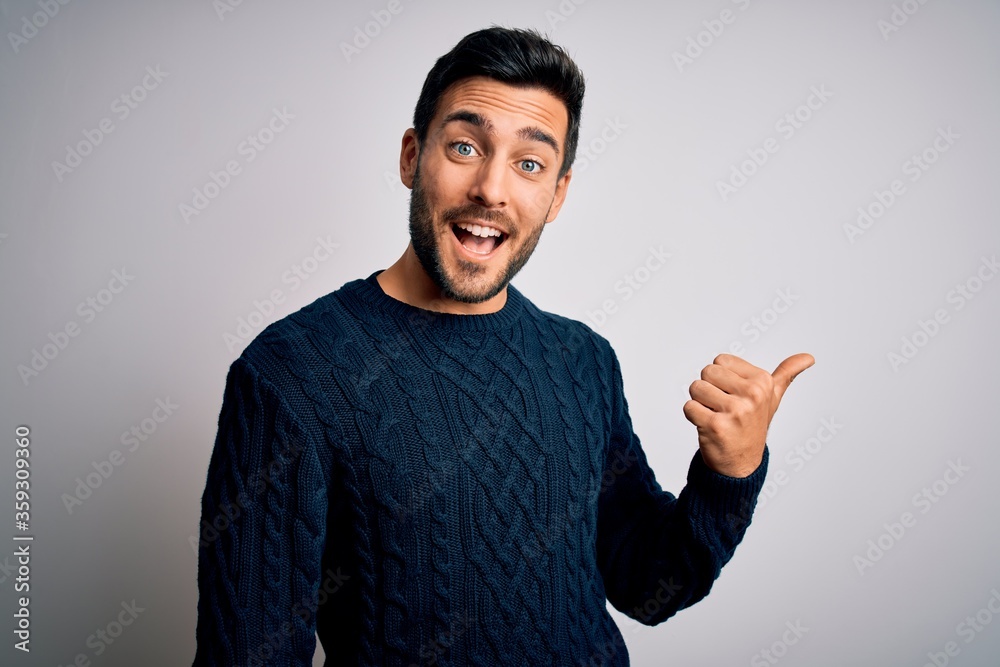 Fototapeta premium Young handsome man with beard wearing casual sweater standing over white background smiling with happy face looking and pointing to the side with thumb up.