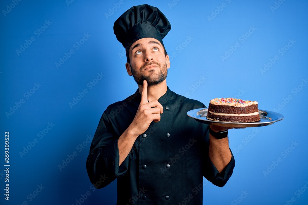 Young handsome cooker man with beard wearing uniform and hat holding tray with cake serious face thinking about question, very confused idea Young handsome cooker man with beard wearing uniform and hat holding tray with cake serious face thinking about question, very confused idea