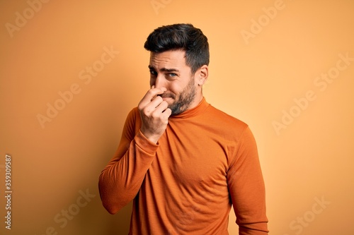 Young handsome man with beard wearing casual sweater standing over yellow background smelling something stinky and disgusting, intolerable smell, holding breath with fingers on nose. Bad smell