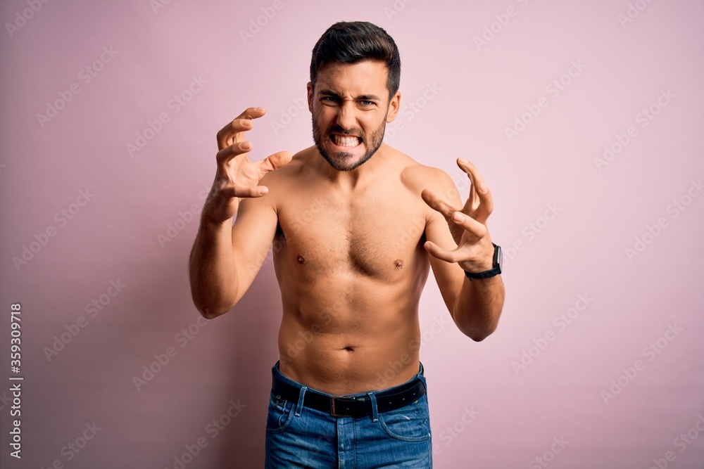 Young handsome strong man with beard shirtless standing over isolated pink background Shouting frustrated with rage, hands trying to strangle, yelling mad