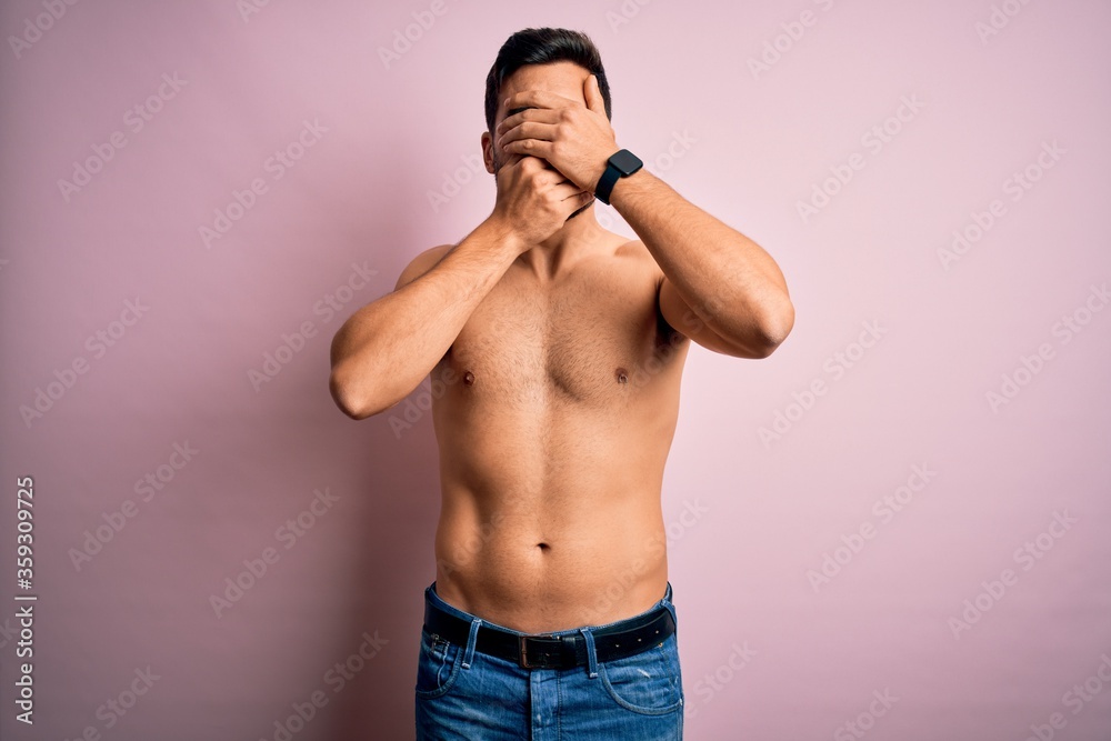 Young handsome strong man with beard shirtless standing over isolated pink background Covering eyes and mouth with hands, surprised and shocked. Hiding emotion