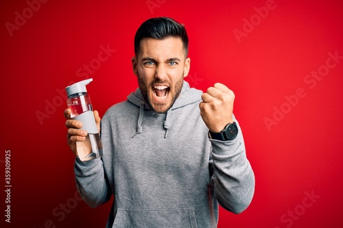 Young handsome sportsman drinking bottle of water to refeshment over red background annoyed and frustrated shouting with anger, crazy and yelling with raised hand, anger concept photo