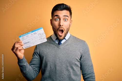 Young handsome tourist man holding vacation boarding plass over yellow isolated background scared in shock with a surprise face, afraid and excited with fear expression photo