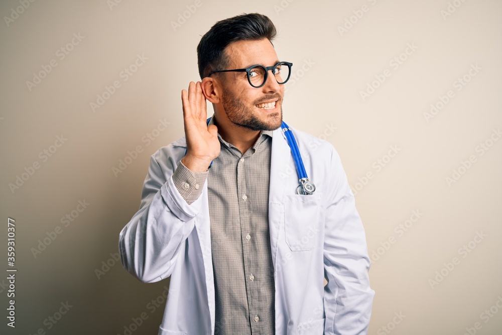 Young doctor man wearing glasses, medical white robe and stethoscope over isolated background smiling with hand over ear listening an hearing to rumor or gossip. Deafness concept. Young doctor man wearing glasses, medical white robe and stethoscope over isolated background smiling with hand over ear listening an hearing to rumor or gossip. Deafness concept.