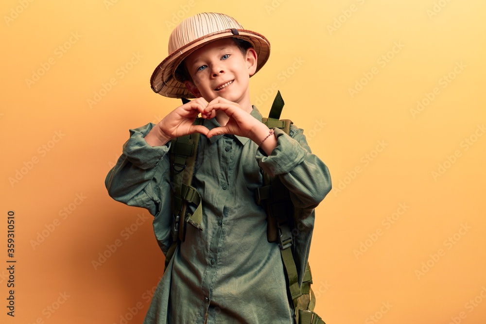Cute blond kid wearing explorer hat and backpack smiling in love doing heart symbol shape with hands. romantic concept. Cute blond kid wearing explorer hat and backpack smiling in love doing heart symbol shape with hands. romantic concept.