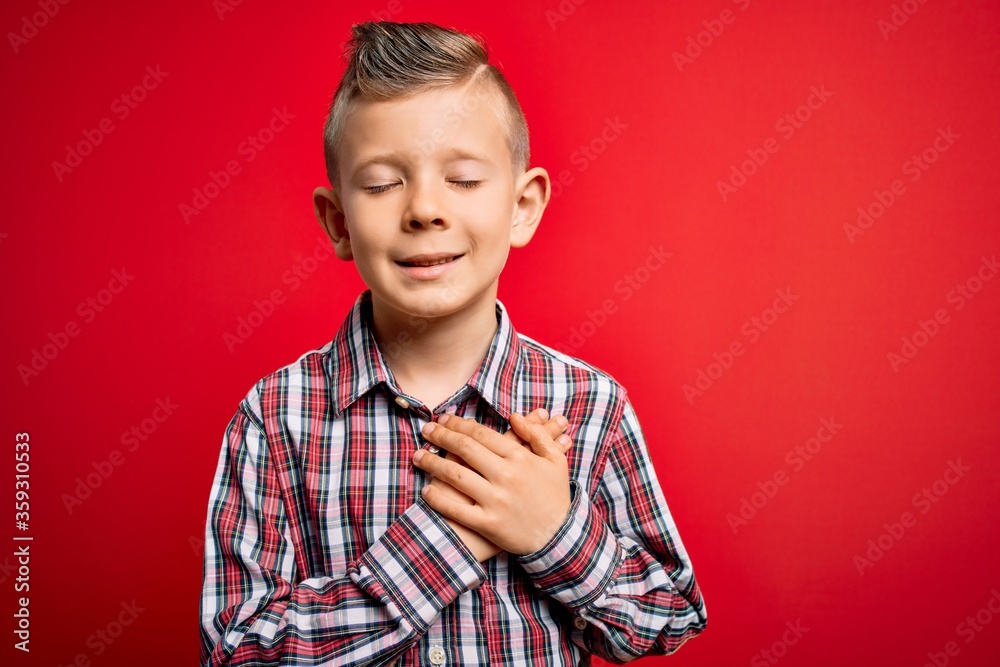 Young little caucasian kid with blue eyes standing wearing elegant shirt over red background smiling with hands on chest with closed eyes and grateful gesture on face. Health concept. Young little caucasian kid with blue eyes standing wearing elegant shirt over red background smiling with hands on chest with closed eyes and grateful gesture on face. Health concept.