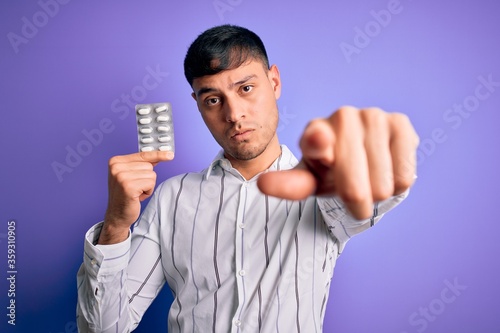 Young hispanic man holding pharmaceutical antibiotics pills over purple isolated background pointing with finger to the camera and to you, hand sign, positive and confident gesture from the front