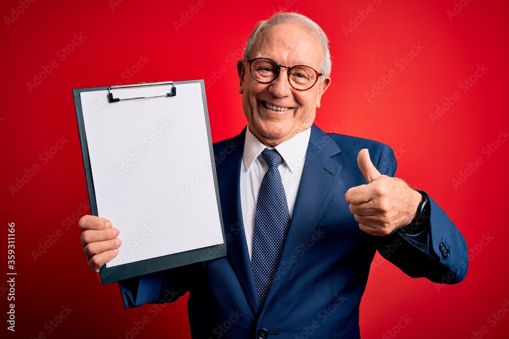 Senior grey haired business man holding clipboard over red background happy with big smile doing ok sign, thumb up with fingers, excellent sign Senior grey haired business man holding clipboard over red background happy with big smile doing ok sign, thumb up with fingers, excellent sign