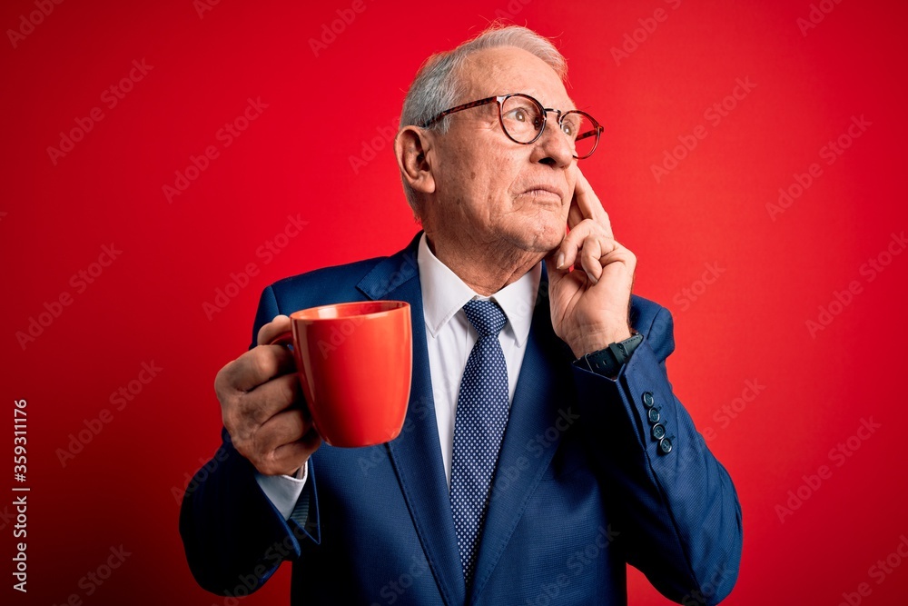 Senior grey haired business man drinking a hot cup of coffee over red background serious face thinking about question, very confused idea Senior grey haired business man drinking a hot cup of coffee over red background serious face thinking about question, very confused idea