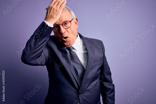 Grey haired senior business man wearing glasses and elegant suit and tie over purple background surprised with hand on head for mistake, remember error. Forgot, bad memory concept.