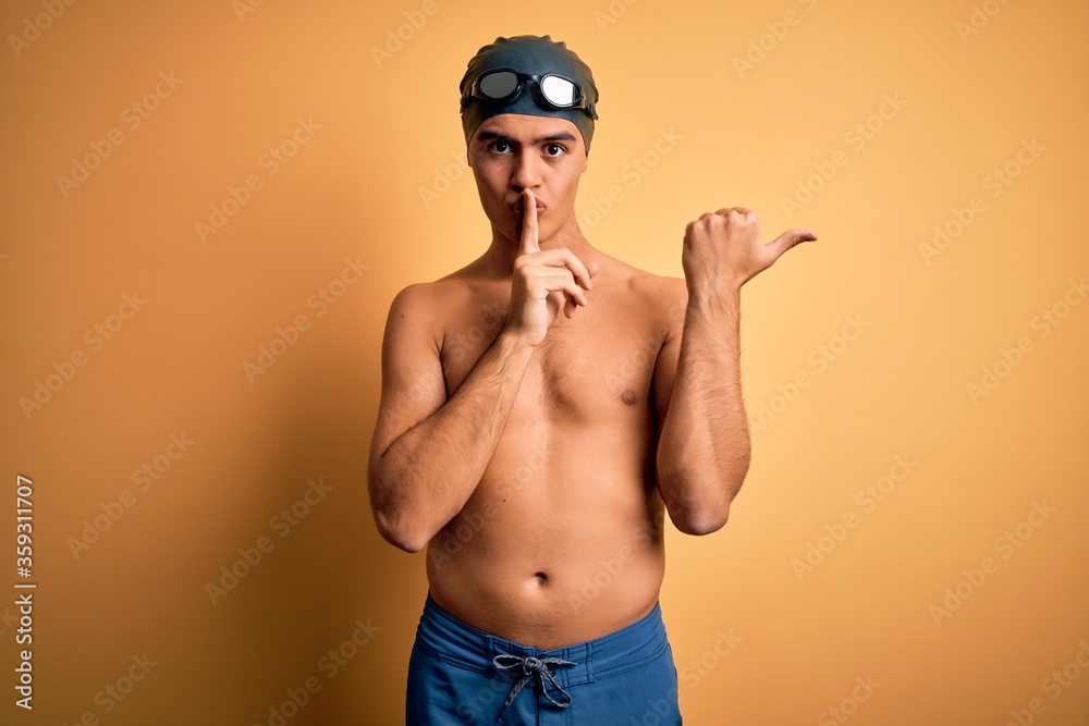 Young handsome man shirtless wearing swimsuit and swim cap over isolated yellow background asking to be quiet with finger on lips pointing with hand to the side. Silence and secret concept.