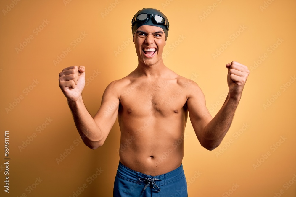 Young handsome man shirtless wearing swimsuit and swim cap over isolated yellow background angry and mad raising fists frustrated and furious while shouting with anger. Rage and aggressive concept.