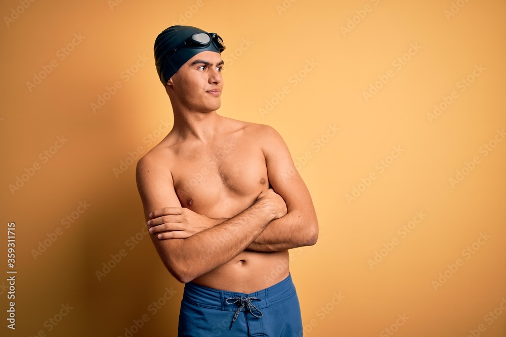 Young handsome man shirtless wearing swimsuit and swim cap over isolated yellow background looking to the side with arms crossed convinced and confident