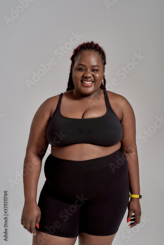 Be comfortable. Plump, plus size african american woman in black sportswear laughing while standing in studio over grey background. Concept of sport, healthy lifestyle, body positive, equality © Svitlana