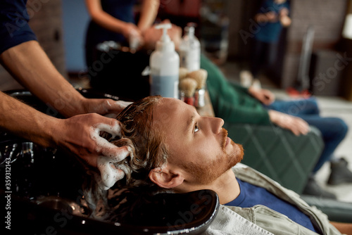 Side view of a young handsome redhead man leaning on the sink while barber washing his hair before a haircut
