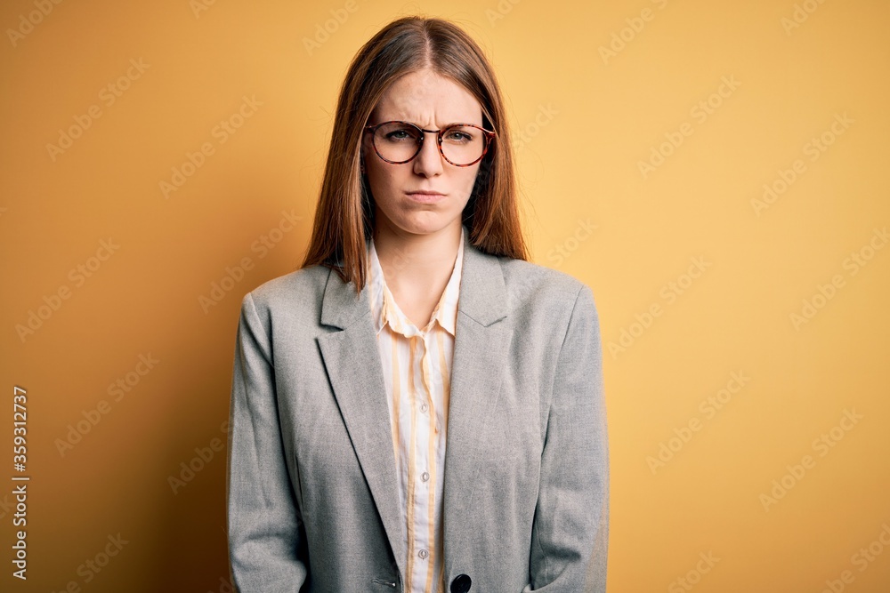 Young beautiful redhead woman wearing jacket and glasses over isolated yellow background skeptic and nervous, frowning upset because of problem. Negative person.