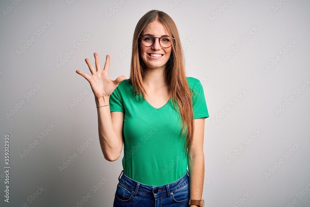 Young beautiful redhead woman wearing casual green t-shirt and glasses over white background showing and pointing up with fingers number five while smiling confident and happy.