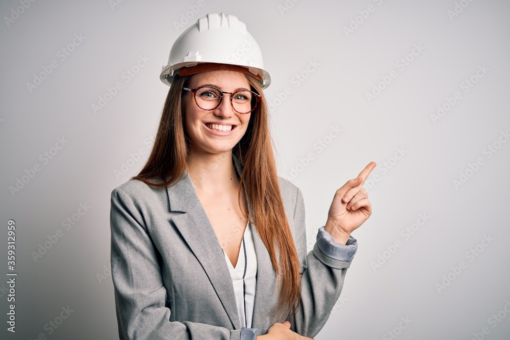 Young beautiful redhead architect woman wearing security helmet over white background with a big smile on face, pointing with hand and finger to the side looking at the camera. Young beautiful redhead architect woman wearing security helmet over white background with a big smile on face, pointing with hand and finger to the side looking at the camera.