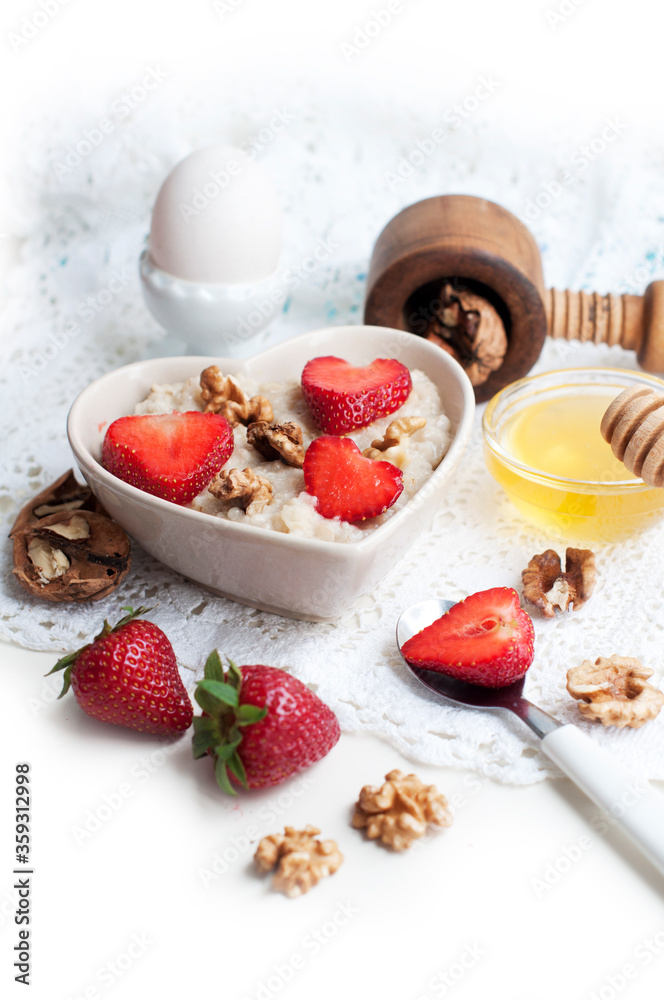 Breakfast with oatmeal with strawberries, honey, walnuts and egg on white background. Breakfast for child. Oatmeal in a white heart-shaped bowl Breakfast with oatmeal with strawberries, honey, walnuts and egg on white background. Breakfast for child. Oatmeal in a white heart-shaped bowl