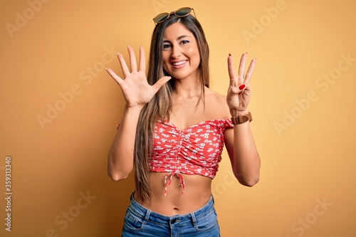 Young beautiful woman wearing summer style and sunglasses over yellow isolated background showing and pointing up with fingers number eight while smiling confident and happy.