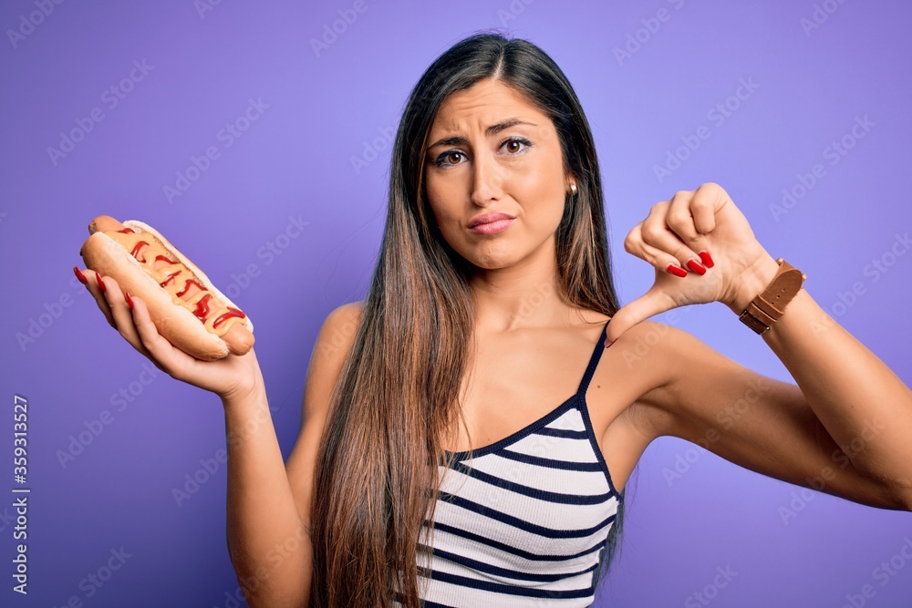 Young woman eating hotdog with ketchup and mustard over purple background with angry face, negative sign showing dislike with thumbs down, rejection concept