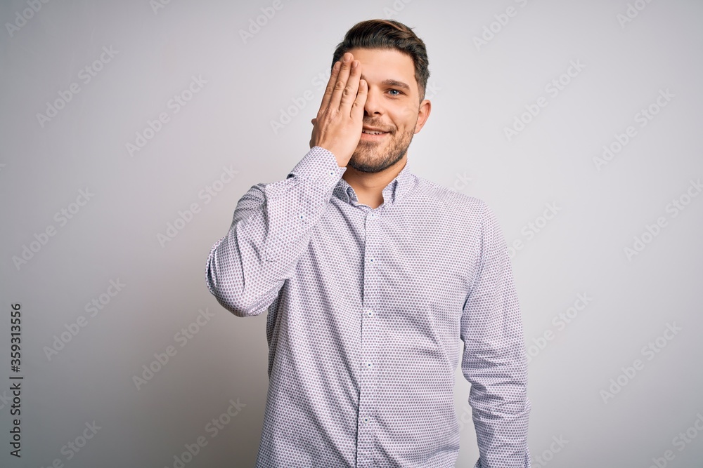 Young business man with blue eyes standing over isolated background covering one eye with hand, confident smile on face and surprise emotion. Young business man with blue eyes standing over isolated background covering one eye with hand, confident smile on face and surprise emotion.