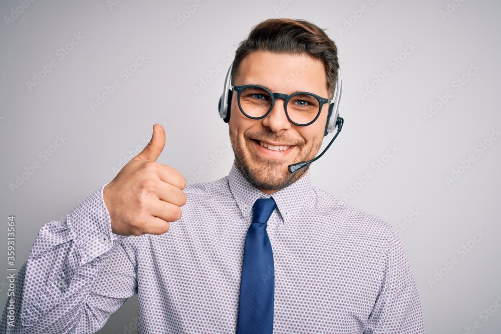 Young call center operator business man with blue eyes wearing glasses and headset doing happy thumbs up gesture with hand. Approving expression looking at the camera showing success. Young call center operator business man with blue eyes wearing glasses and headset doing happy thumbs up gesture with hand. Approving expression looking at the camera showing success.