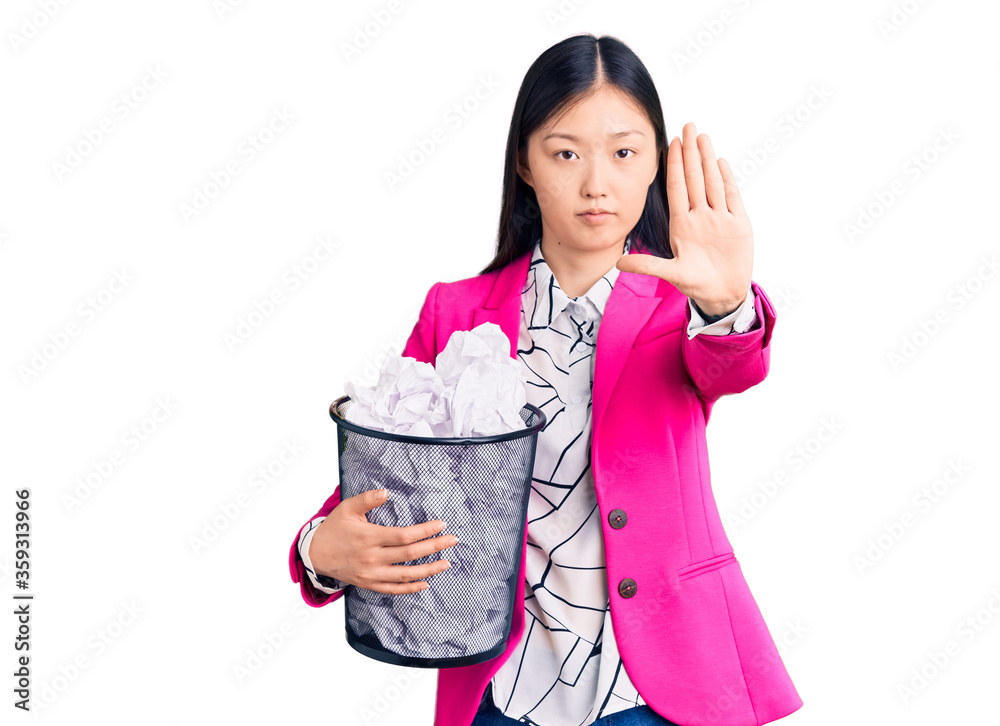 Young beautiful chinese woman holding paper bin full of crumpled papers with open hand doing stop sign with serious and confident expression, defense gesture Young beautiful chinese woman holding paper bin full of crumpled papers with open hand doing stop sign with serious and confident expression, defense gesture