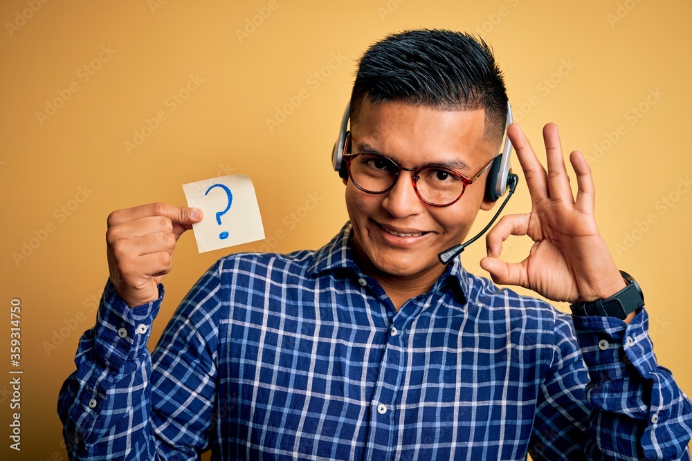 Young latin call center agent man holding question mark reminder paper using headset doing ok sign with fingers, excellent symbol Young latin call center agent man holding question mark reminder paper using headset doing ok sign with fingers, excellent symbol