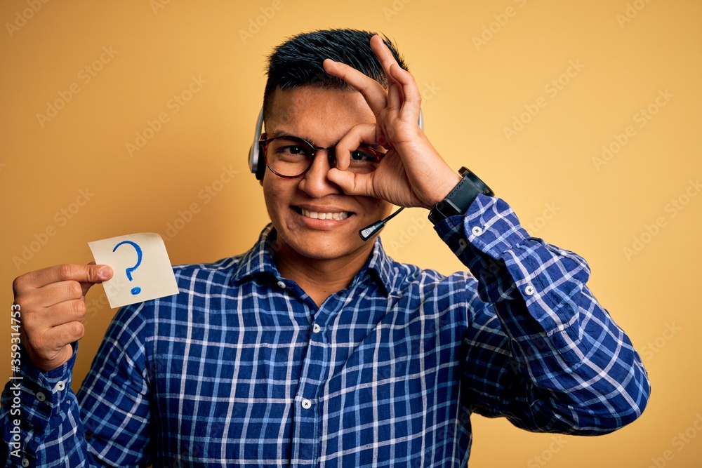 Young latin call center agent man holding question mark reminder paper using headset with happy face smiling doing ok sign with hand on eye looking through fingers Young latin call center agent man holding question mark reminder paper using headset with happy face smiling doing ok sign with hand on eye looking through fingers