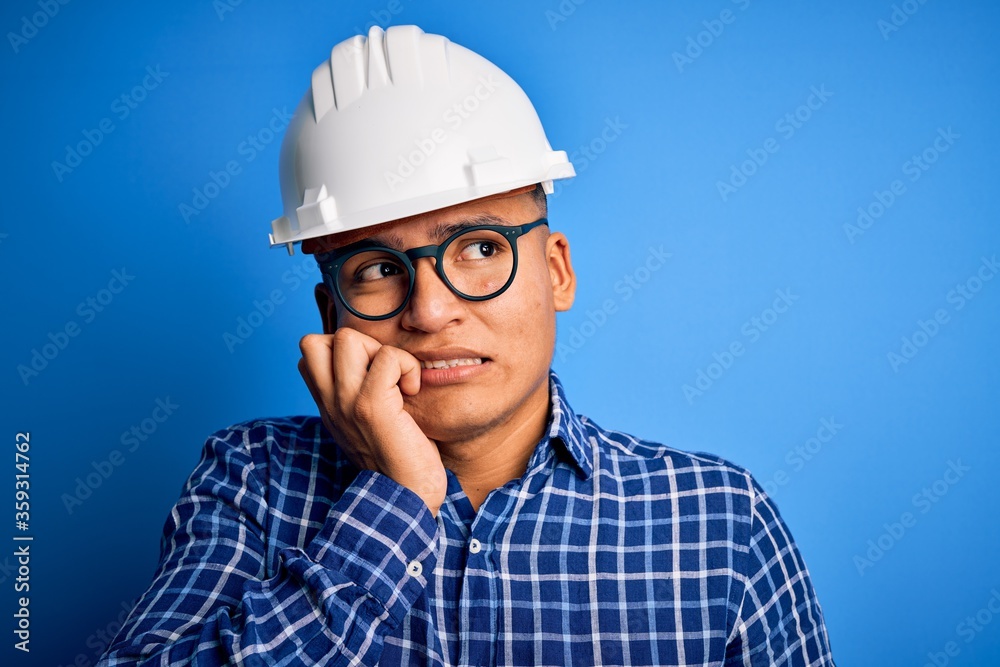 Young handsome engineer latin man wearing safety helmet over isolated blue background looking stressed and nervous with hands on mouth biting nails. Anxiety problem.