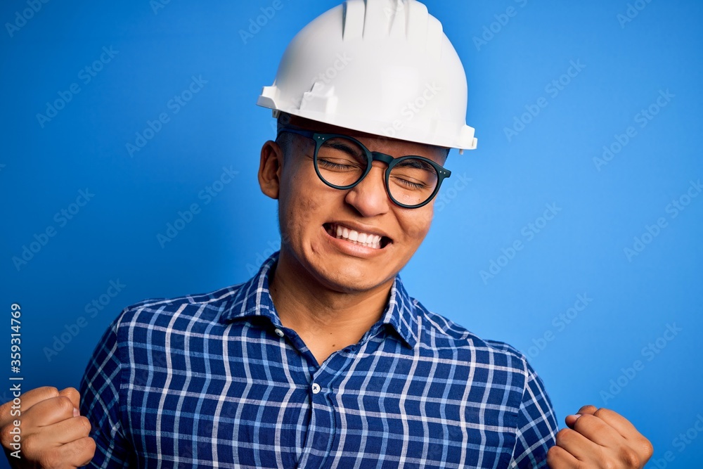 Young handsome engineer latin man wearing safety helmet over isolated blue background very happy and excited doing winner gesture with arms raised, smiling and screaming for success. Celebration Young handsome engineer latin man wearing safety helmet over isolated blue background very happy and excited doing winner gesture with arms raised, smiling and screaming for success. Celebration