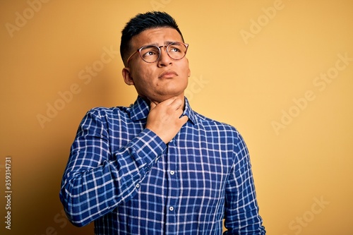 Young handsome latin man wearing casual shirt and glasses over yellow background Touching painful neck, sore throat for flu, clod and infection