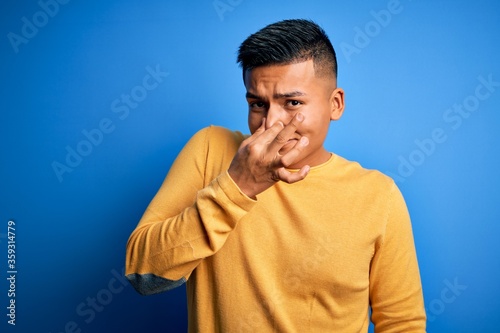 Young handsome latin man wearing yellow casual sweater over isolated blue background smelling something stinky and disgusting, intolerable smell, holding breath with fingers on nose. Bad smell