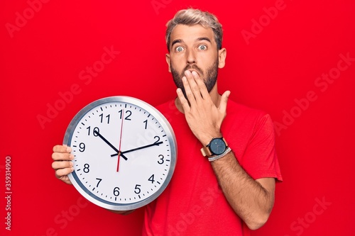 Young handsome blond man with beard doing countdown using big clock over red background covering mouth with hand, shocked and afraid for mistake. Surprised expression