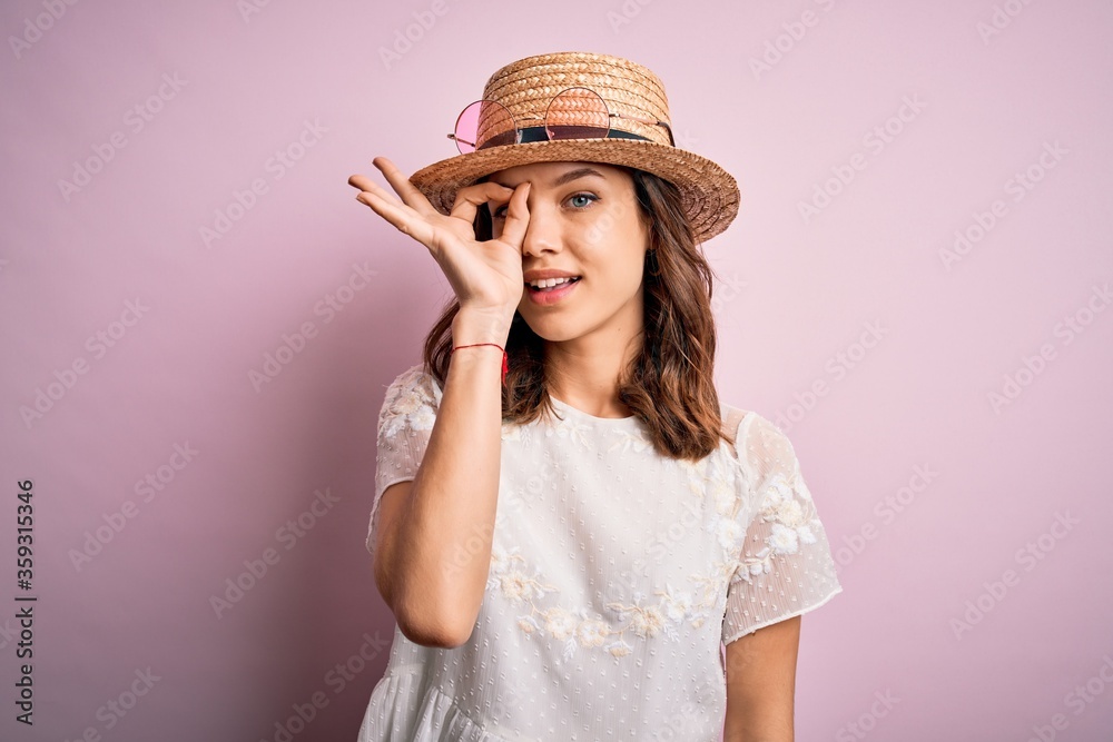 Young beautiful blonde girl wearing summer hat over pink isolated background doing ok gesture with hand smiling, eye looking through fingers with happy face. Young beautiful blonde girl wearing summer hat over pink isolated background doing ok gesture with hand smiling, eye looking through fingers with happy face.