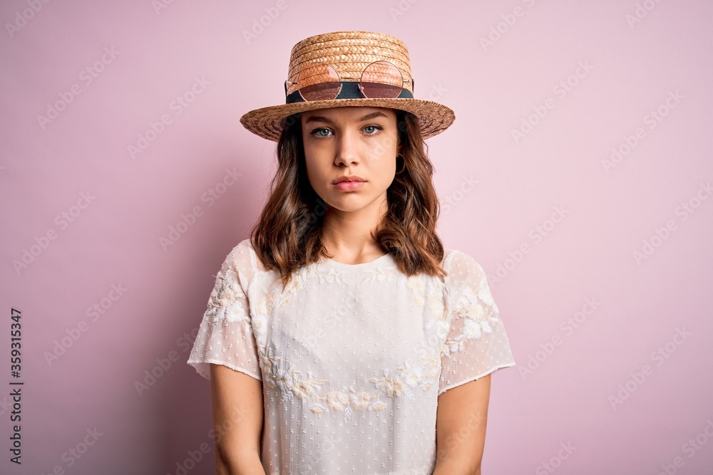 Young beautiful blonde girl wearing summer hat over pink isolated background with serious expression on face. Simple and natural looking at the camera. Young beautiful blonde girl wearing summer hat over pink isolated background with serious expression on face. Simple and natural looking at the camera.