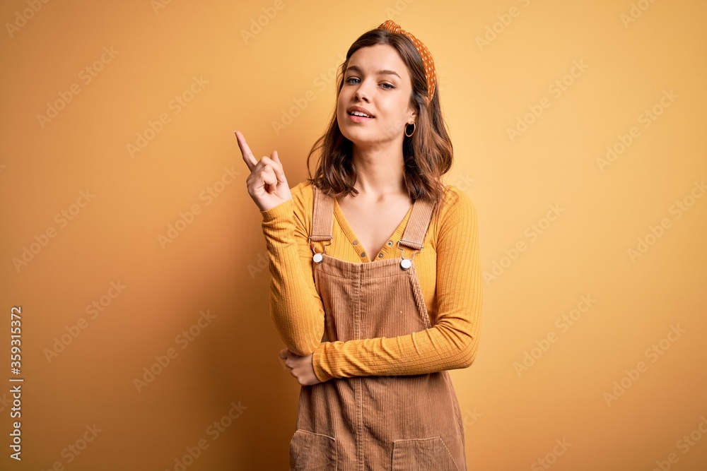 Young beautiful blonde girl wearing overall standing over yellow isolated background with a big smile on face, pointing with hand finger to the side looking at the camera. Young beautiful blonde girl wearing overall standing over yellow isolated background with a big smile on face, pointing with hand finger to the side looking at the camera.