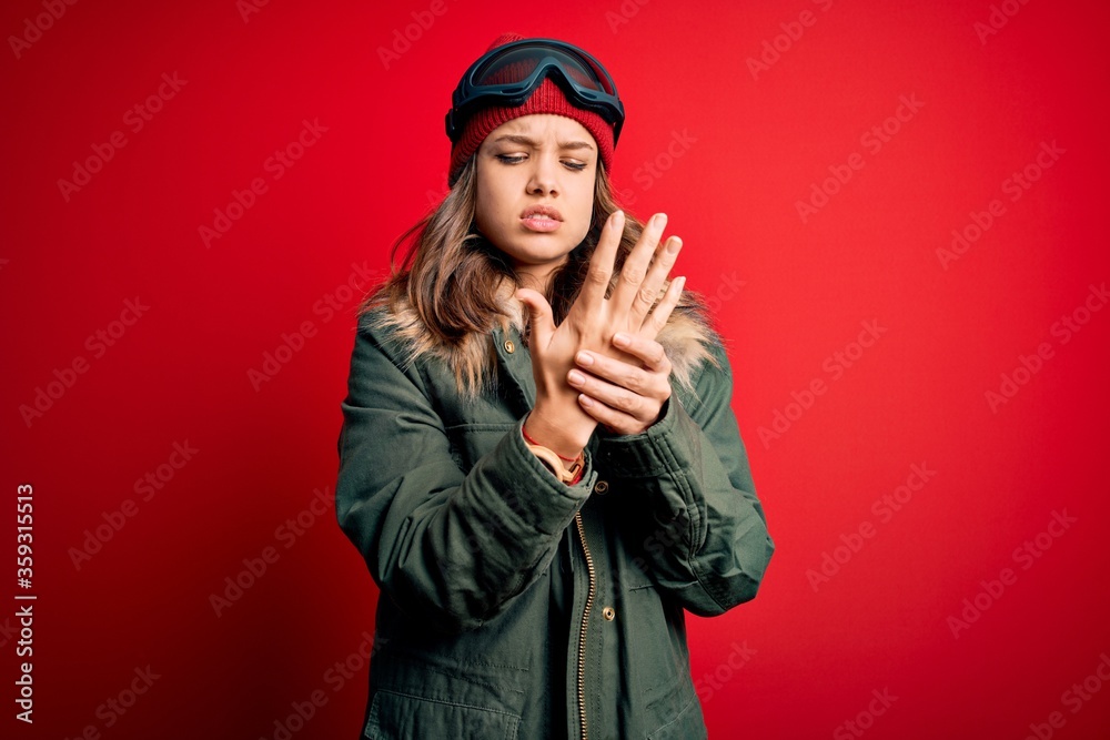 Young blonde girl wearing ski glasses and winter coat for ski weather over red background Suffering pain on hands and fingers, arthritis inflammation Young blonde girl wearing ski glasses and winter coat for ski weather over red background Suffering pain on hands and fingers, arthritis inflammation
