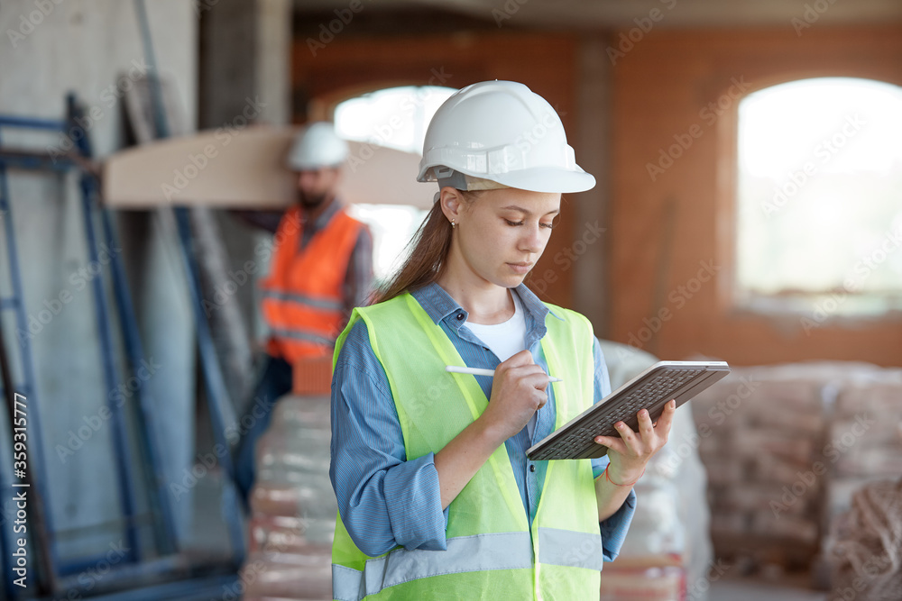 Fototapeta premium A construction industry maintenance engineer is a pretty woman dressed in a uniform and protective helmet with a tablet in her hands against the background of a construction site and a worker