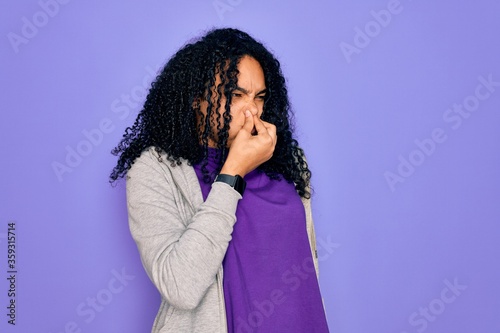 Young african american sporty woman wearing casual sweatshirt over purple background smelling something stinky and disgusting, intolerable smell, holding breath with fingers on nose. Bad smell