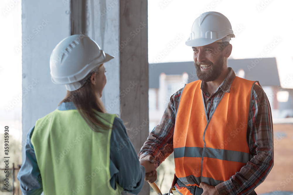 A Caucasian bearded worker in a protective helmet and orange vest shakes hands with a quality control engineer. Business handshake. A Caucasian bearded worker in a protective helmet and orange vest shakes hands with a quality control engineer. Business handshake.