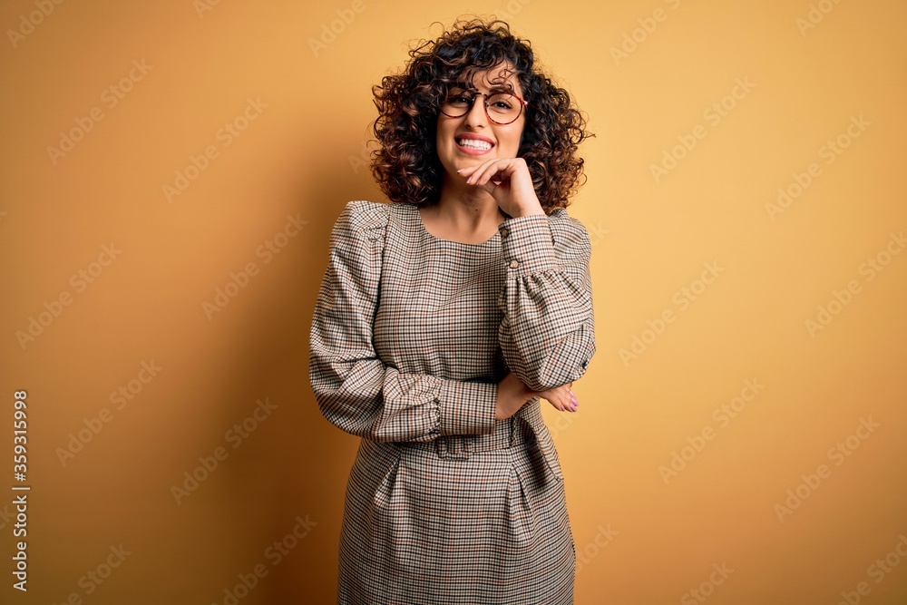 Φωτογραφία Beautiful arab business woman wearing dress and glasses standing over yellow background looking confident at the camera smiling with crossed arms and hand raised on chin