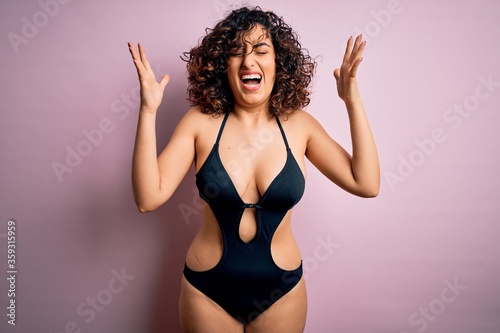 Young beautiful arab woman on vacation wearing swimsuit and sunglasses over pink background celebrating mad and crazy for success with arms raised and closed eyes screaming excited. Winner concept © Krakenimages.com