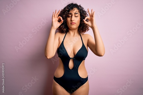 Young beautiful arab woman on vacation wearing swimsuit and sunglasses over pink background relax and smiling with eyes closed doing meditation gesture with fingers. Yoga concept.